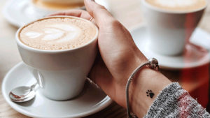 カフェで女性がカフェラテ系の飲み物を持とうとしている写真