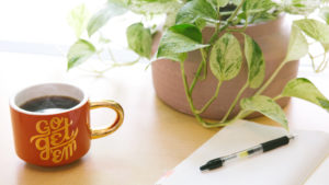 木の机の上に、赤いコップにコーヒーが入っていて、その隣にノートが広げられてペンが置かれている画像
