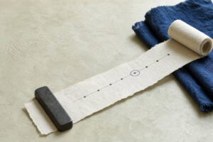 白いしっくいの台の上に広げた細長い和紙に、点と線の年表が描かれ、ひとつの点だけが藍色の丸で囲まれている。端は巻物のように丸まり、藍染めの布と鉄の文鎮が添えられた静物